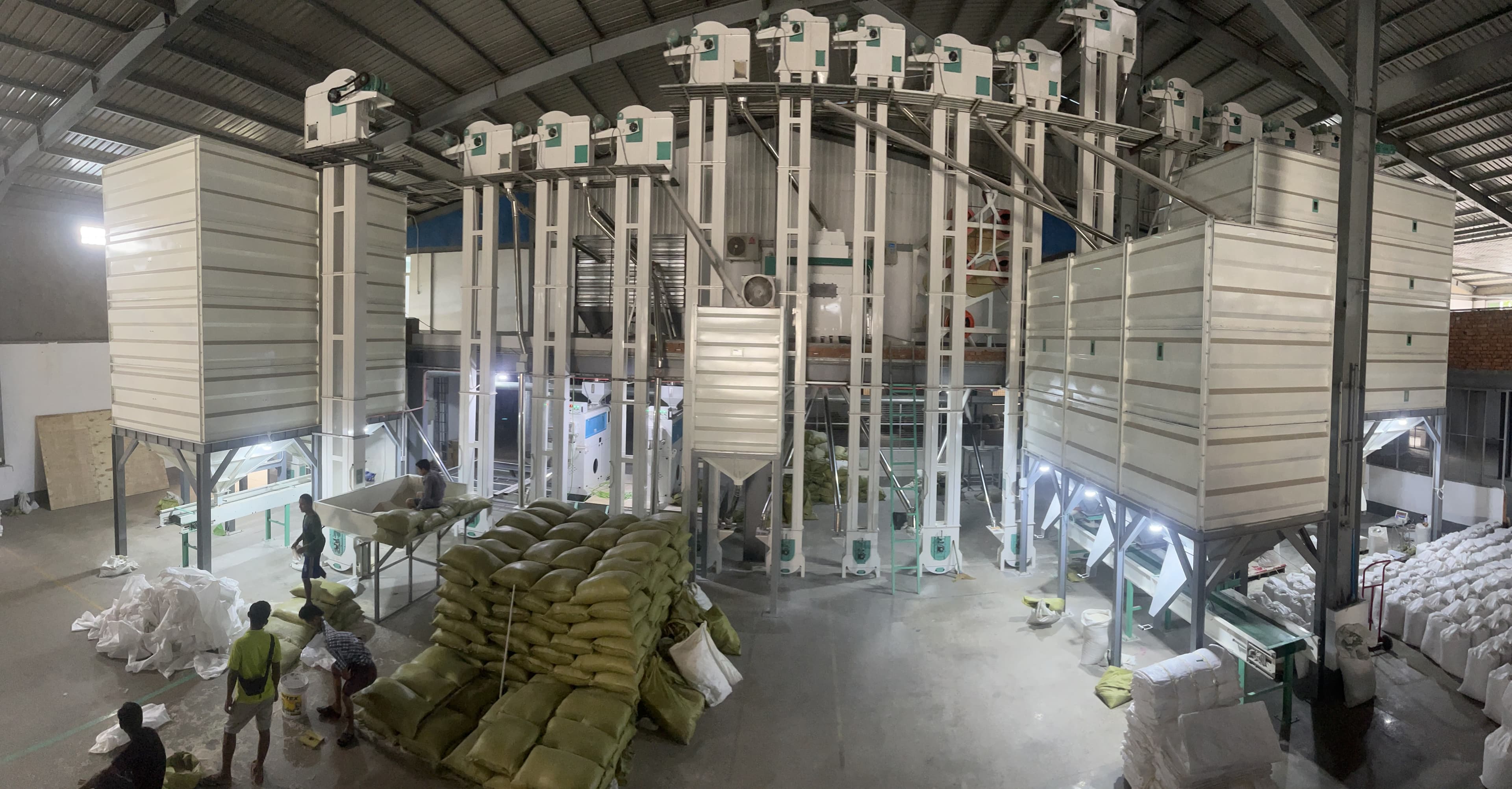 Large industrial grain processing facility with tall white machinery and workers handling stacks of sacks.