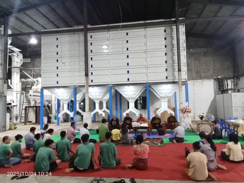 People sitting on a red carpet facing monks before large white industrial silos.